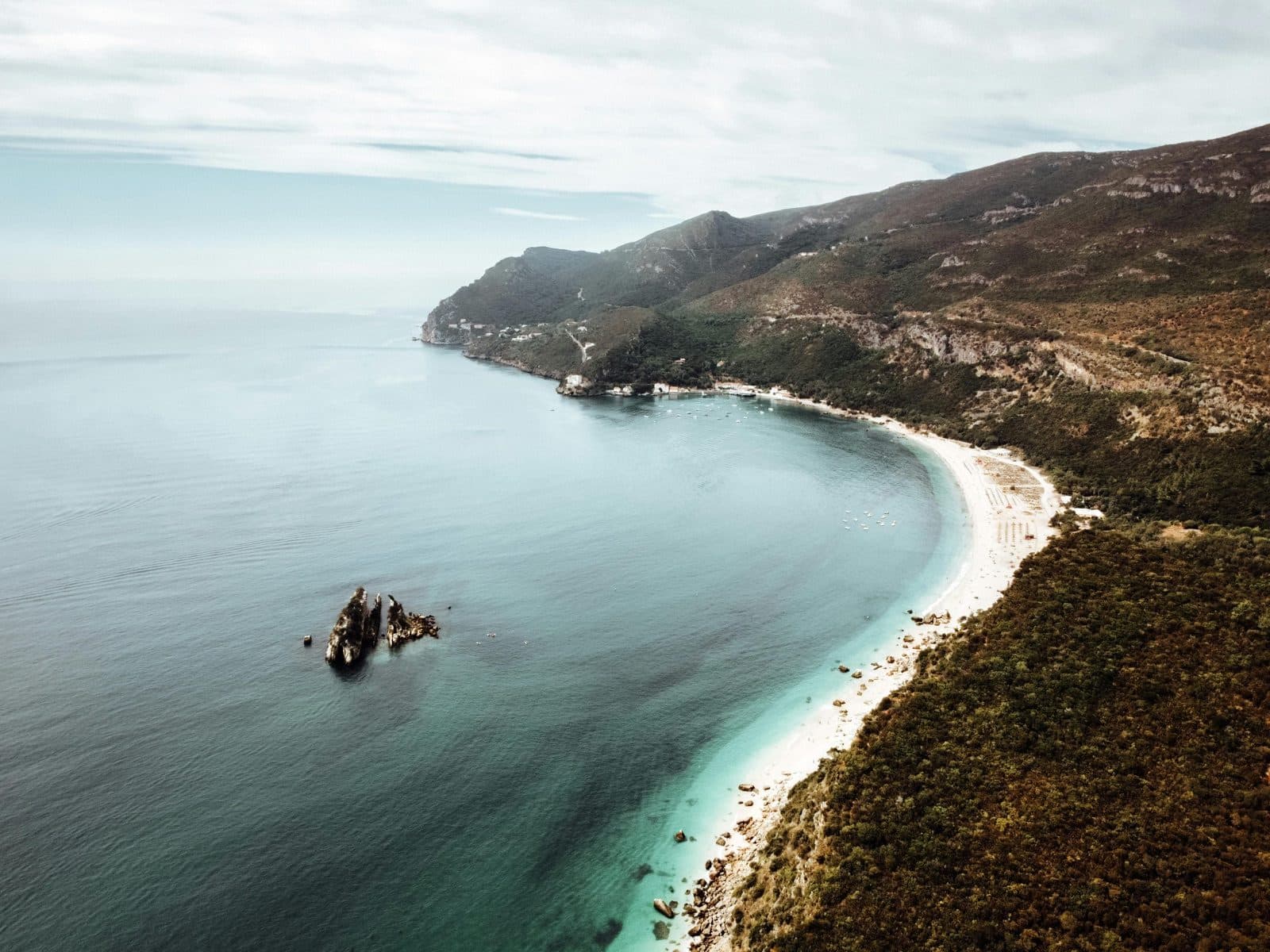 Aerial view of the Arrábida coastline — turquoise water, pine-covered cliffs, white sand beach