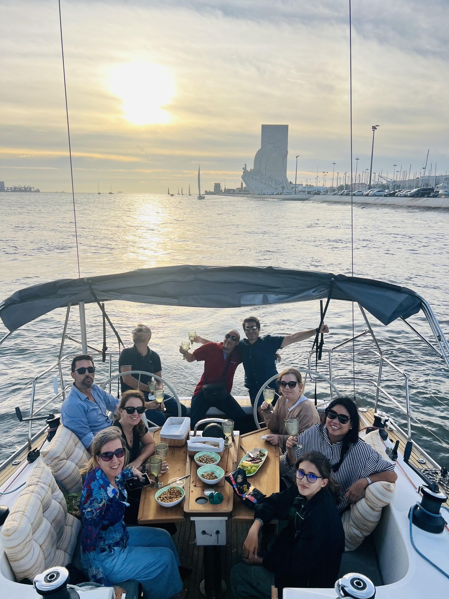 Friends gathered around the cockpit table at sunset on the Tagus, Padrão dos Descobrimentos in the distance